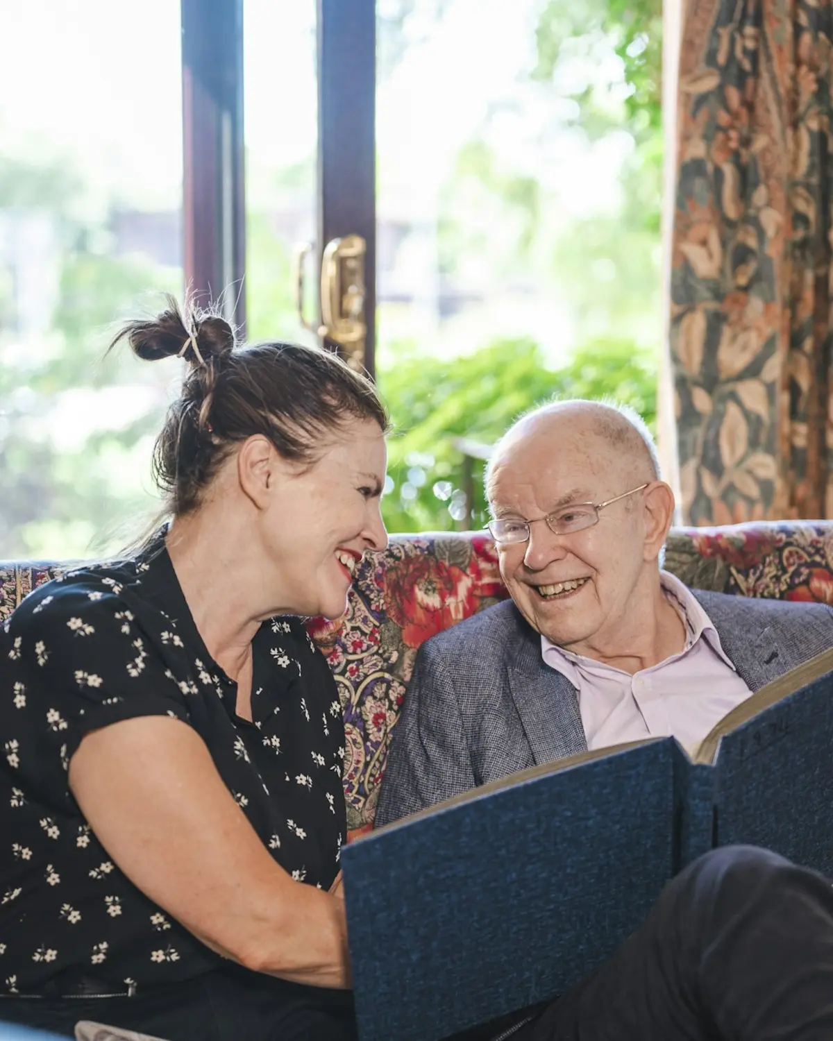 Een oudere man en een jongere vrouw bekijken thuis samen een fotoalbum en lachen.